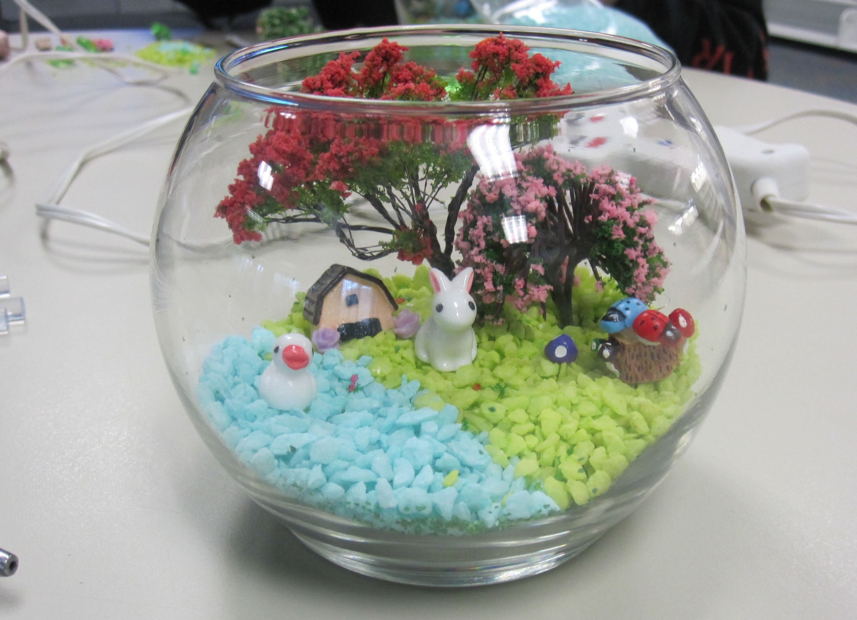 glass bowl terrarium holding colored rocks and sand, small trees, duck figurine, and rabbit figurine