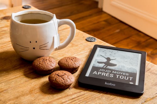 An e-reader sits next to 3 cookies & a cat mug filled with coffee