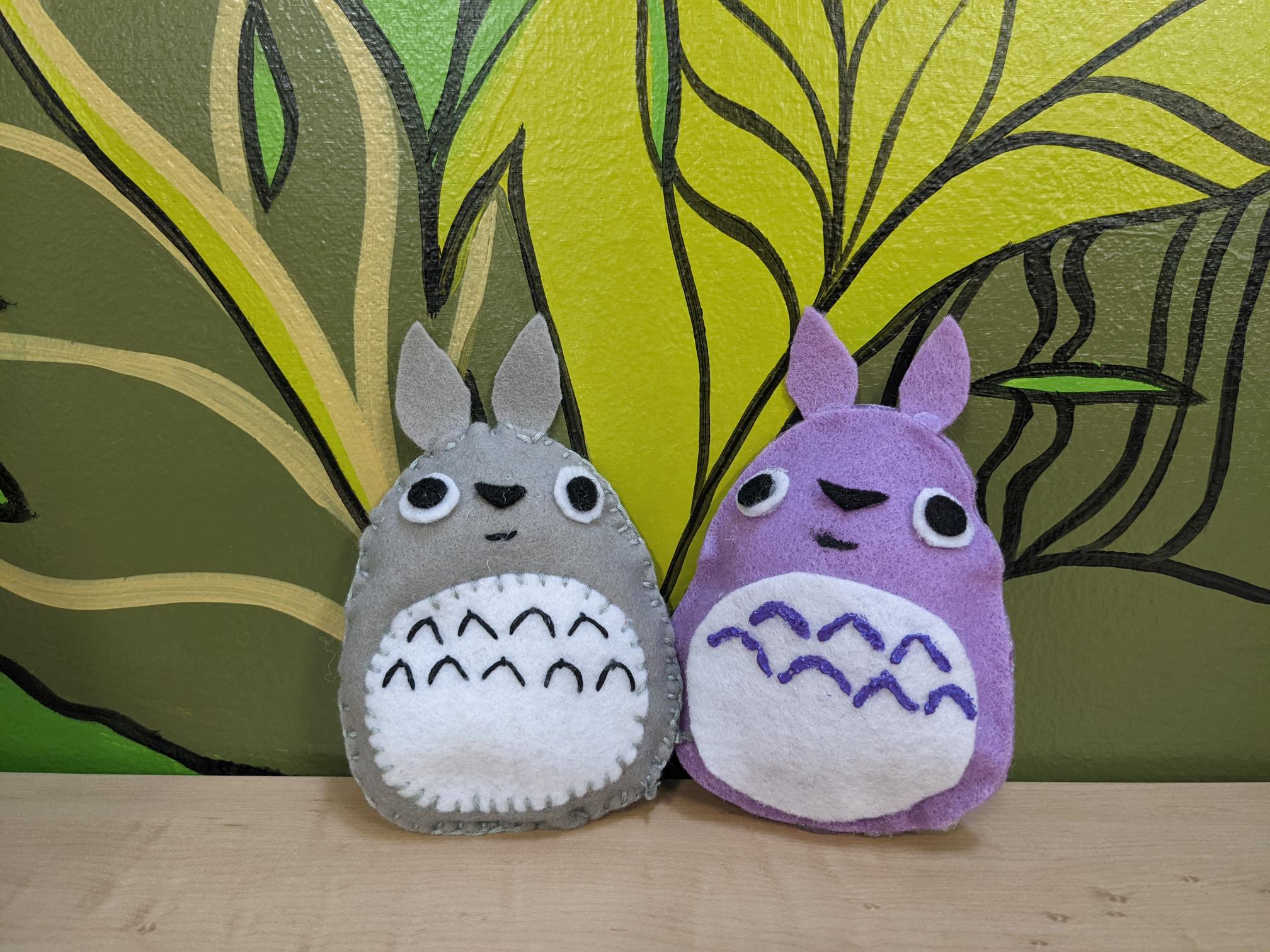 Gray Totoro feltie and lilac colored Totoro feltie against a green leafy background