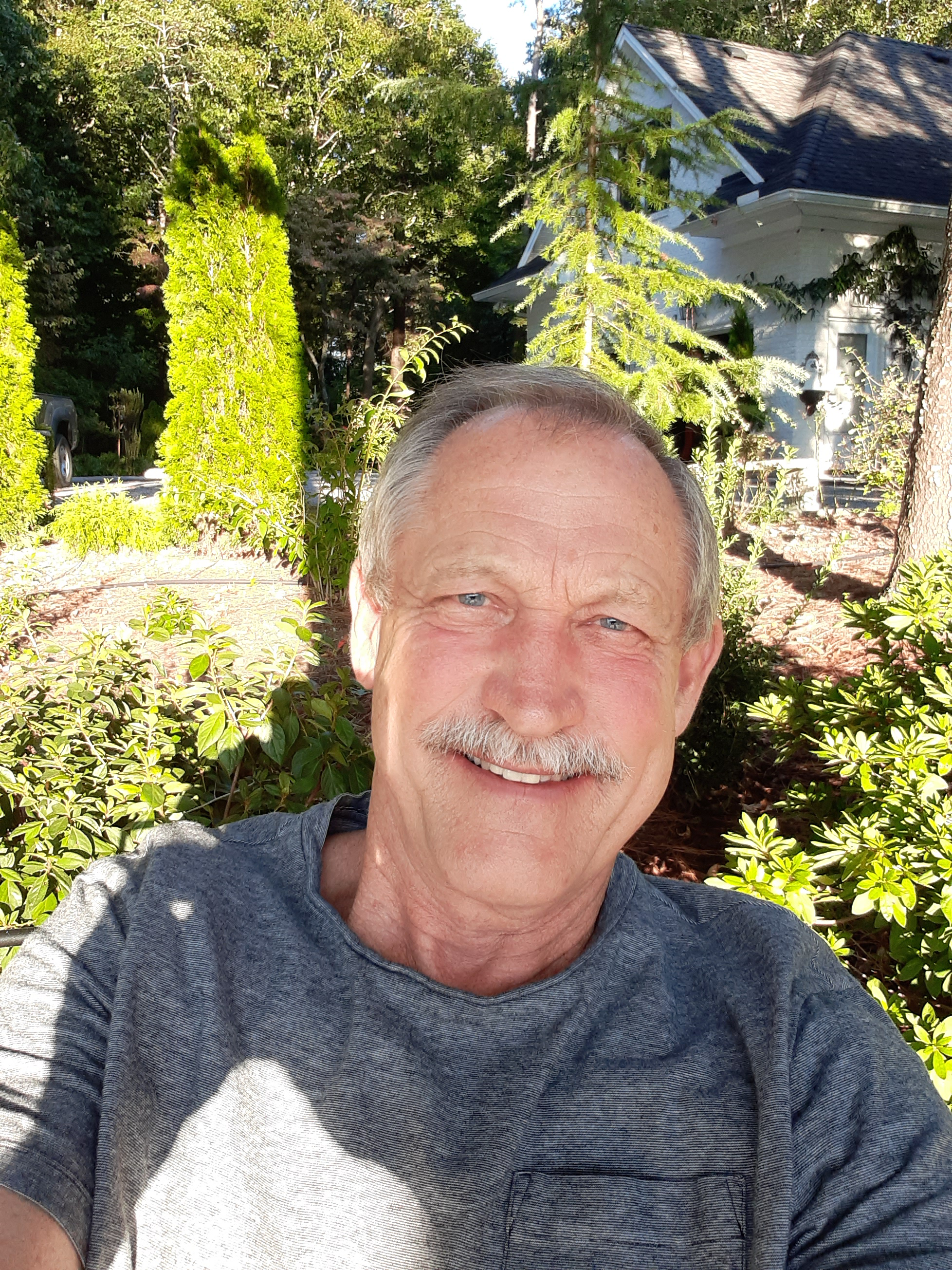 Photo of smiling man with a moustache wearing a gray shirt; a house and trees are in the background
