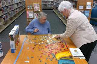 Two women working together to put together a puzzle. One woman is sitting, the other is standing up. 