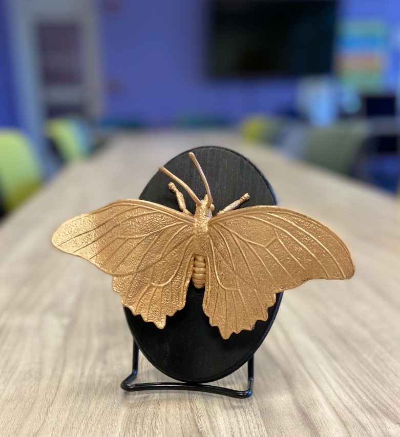 A gilded butterfly on a black plaque. The background is a conference room. 