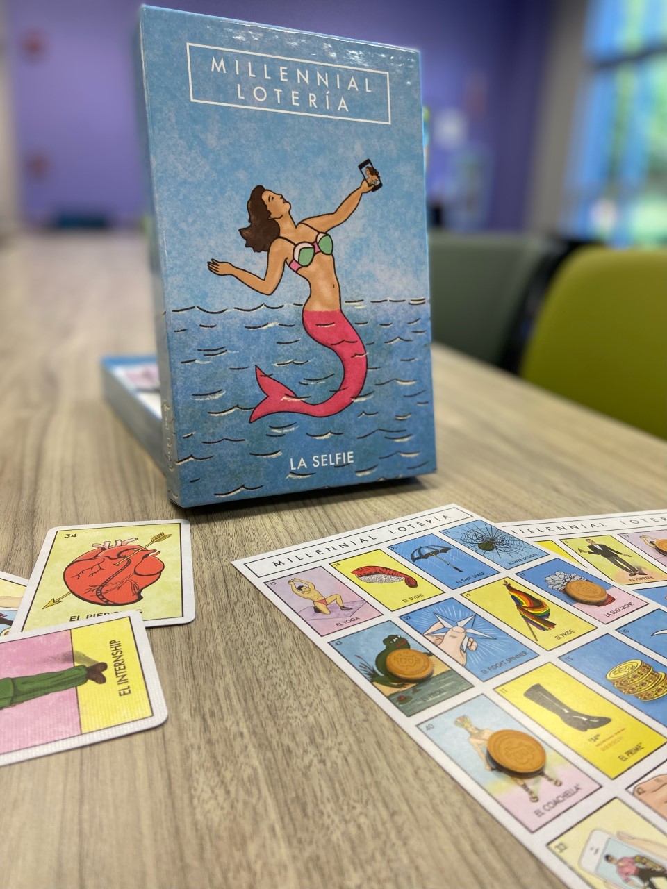 It shows the game Millennial Loteria in the back, with playing cards and boards in the foreground.