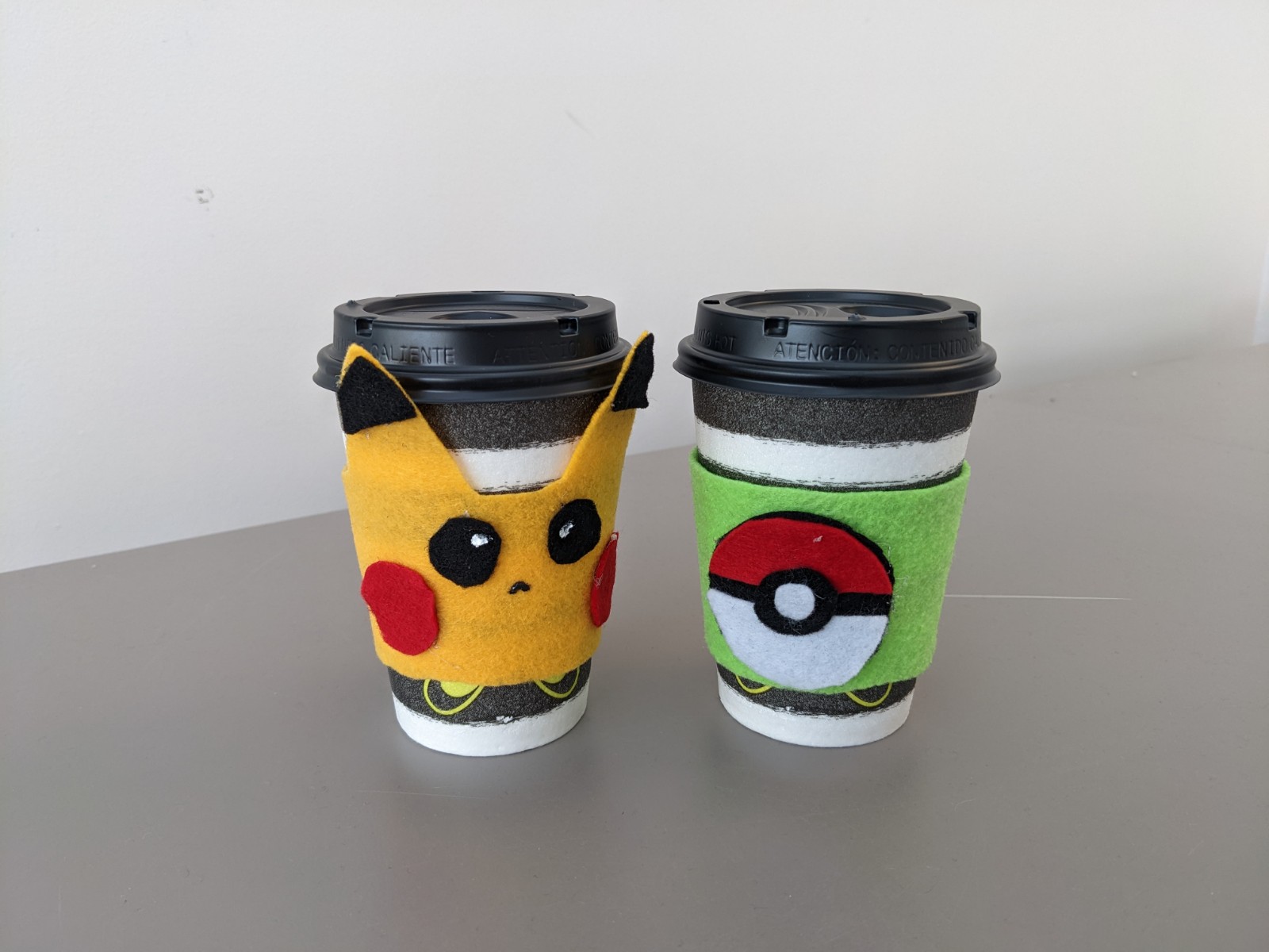 Pikachu face coffee cozy on disposable coffee cup and light green coffee cozy with Pokeball on second disposable coffee cup