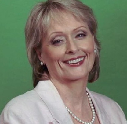 Photo of a woman with short blond hair and a big smile. She is wearing a pale pink jacket and a strand of pearls.