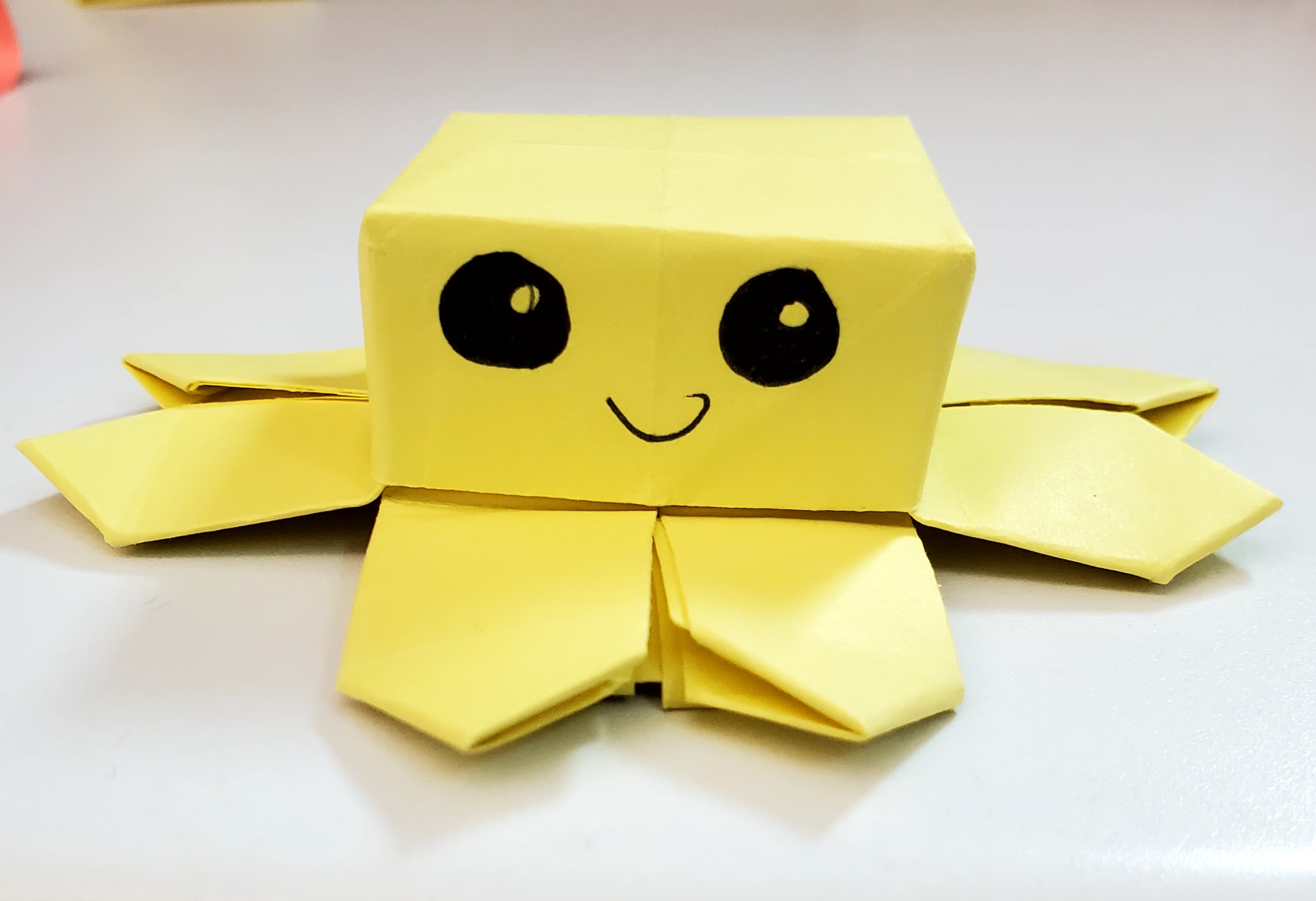 A yellow origami octopus fidget. The head is square shaped and the little critter has big eyes and a smile.