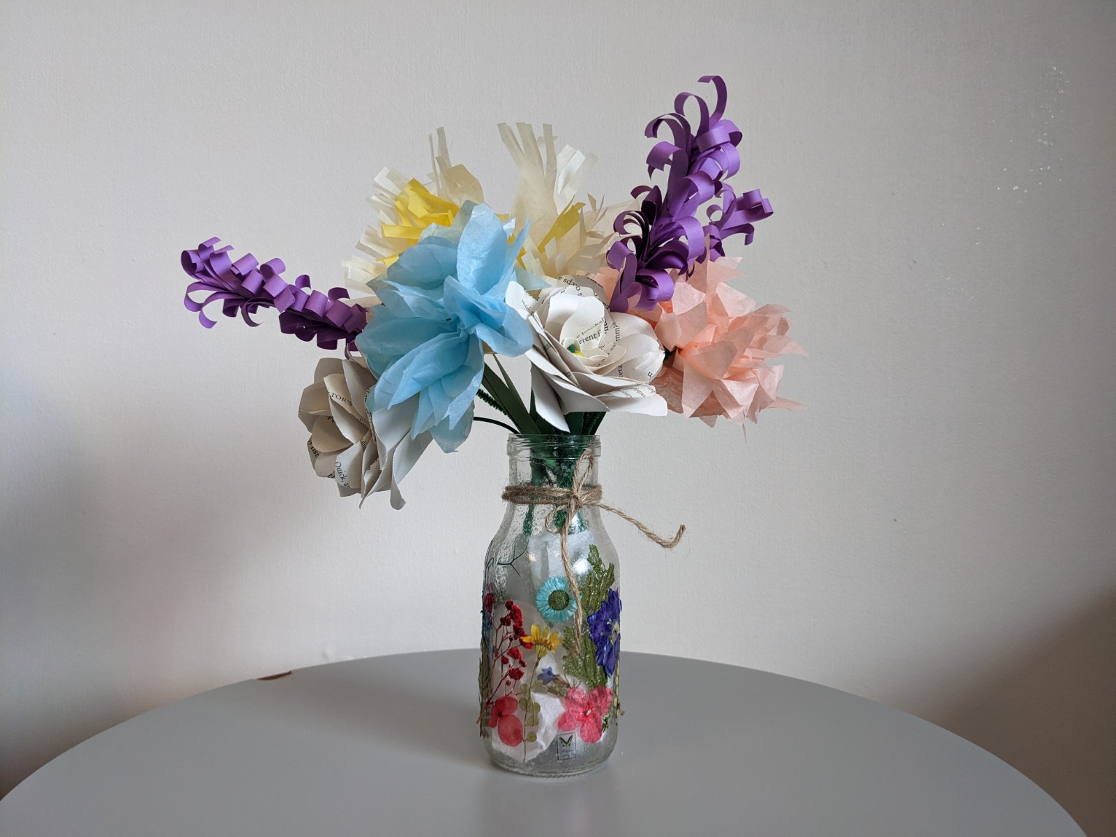 decoupage jar with pressed flowers holding tissue paper flowers in peach, yellow, and light blue, white and book paper roses, and purple paper hyacinth