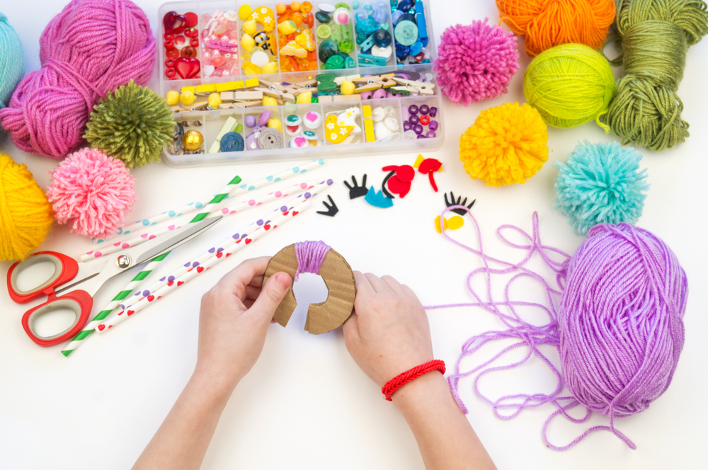 Hands are shown wrapping yarn around a cardboard template in order to make a yarn pom pom. Yarn and other craft materials are in the background on a table.