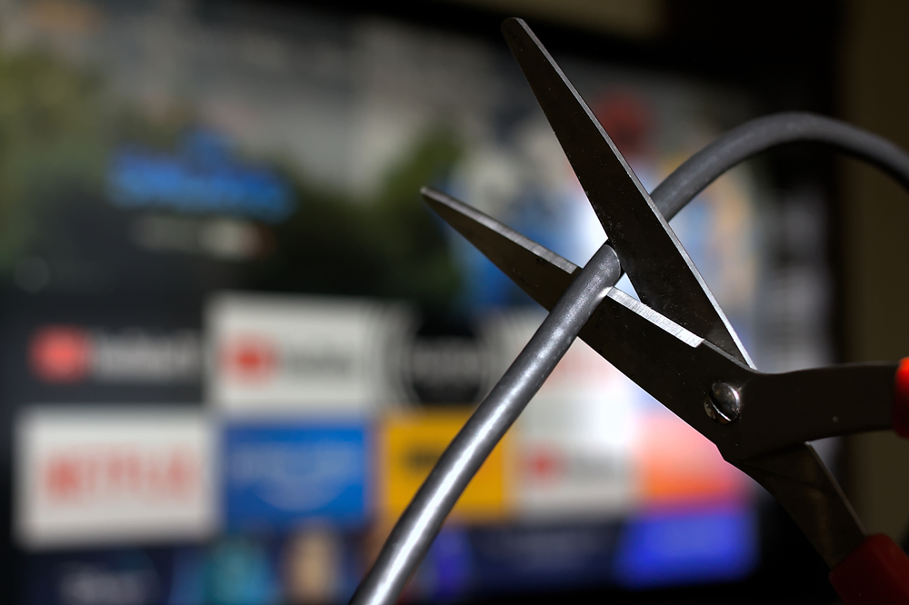 Scissors cutting a cable cord; blurry screen in background showing streaming channels