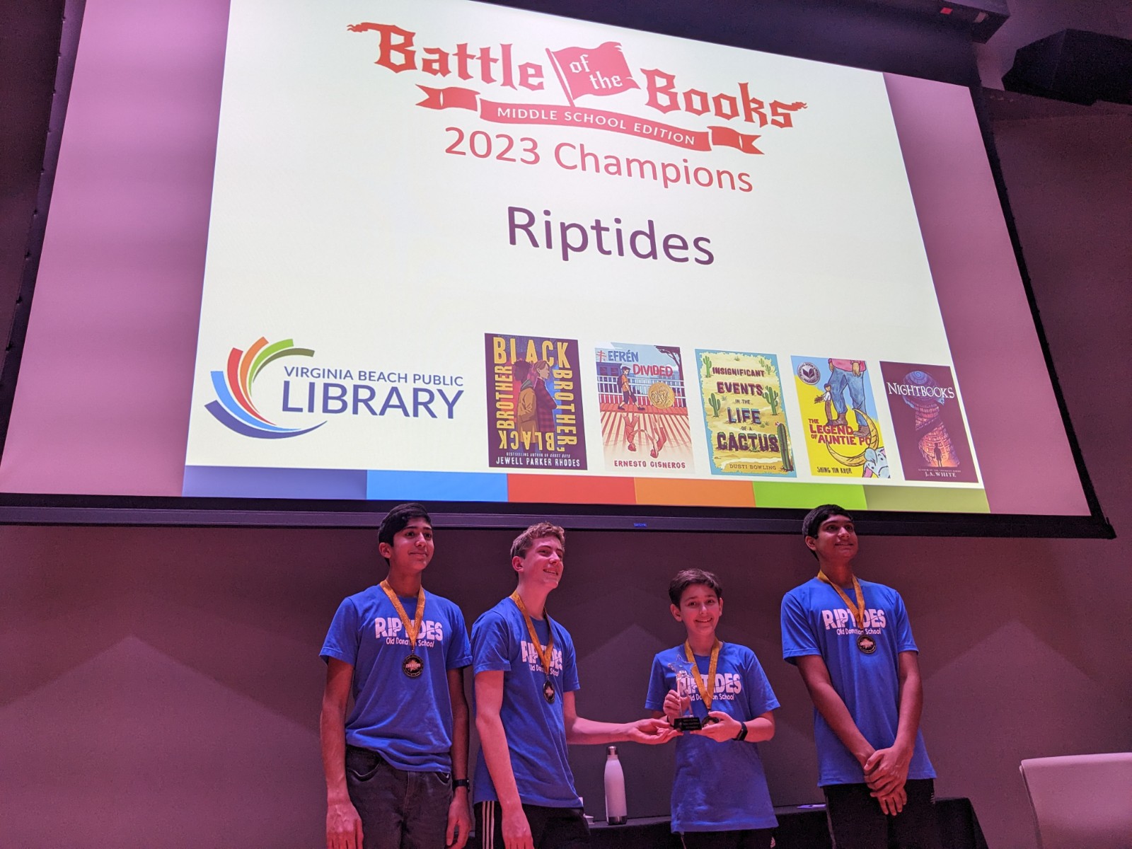 4 teens in blue t shirts and a projection screen saying Battle of the Books Winners: Riptides