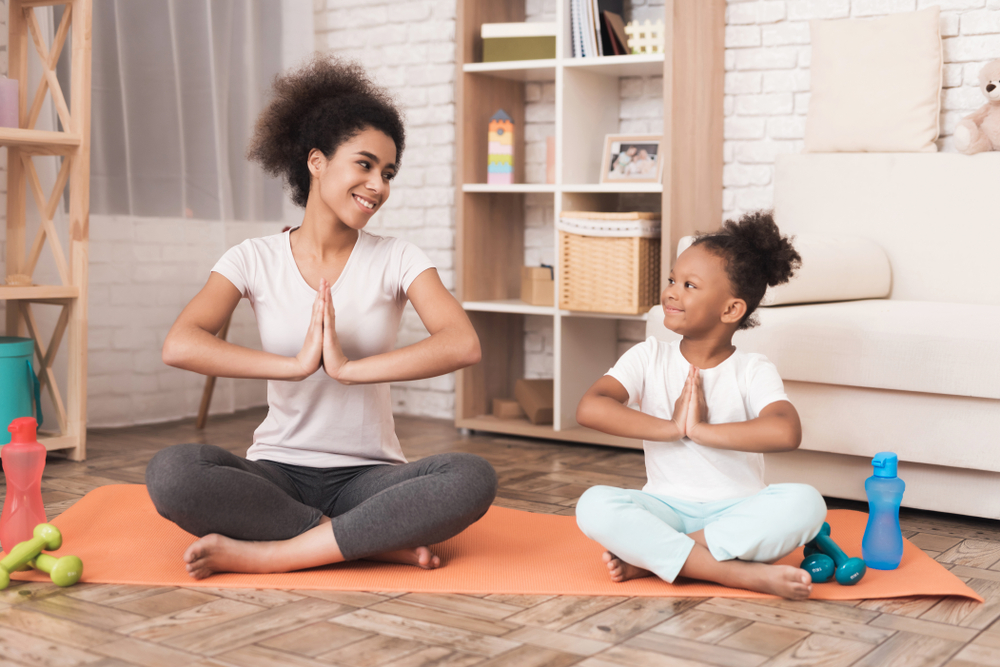 Mom and daughter are doing yoga at home. They are sitting on a rug, next to them are dumbbells