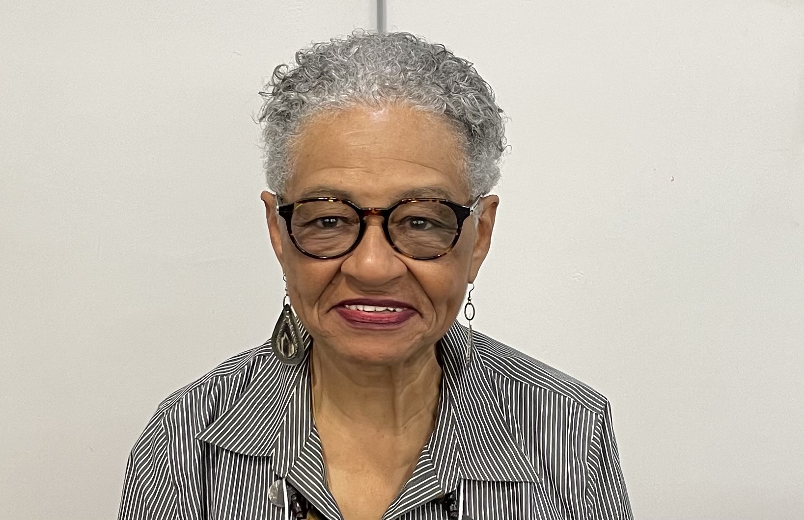 Photo of African American woman with close cropped gray hair & black framed glasses. She is smiling.