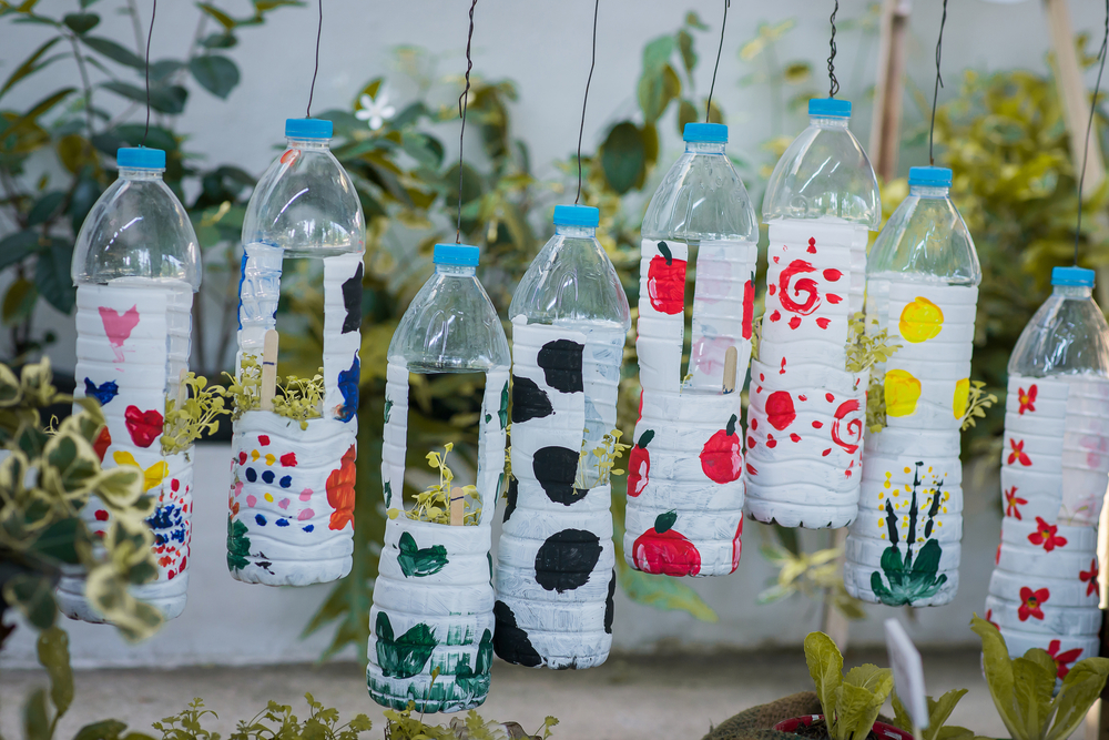 Plastic bottles painted in different colors hanging against a wall with green plants.