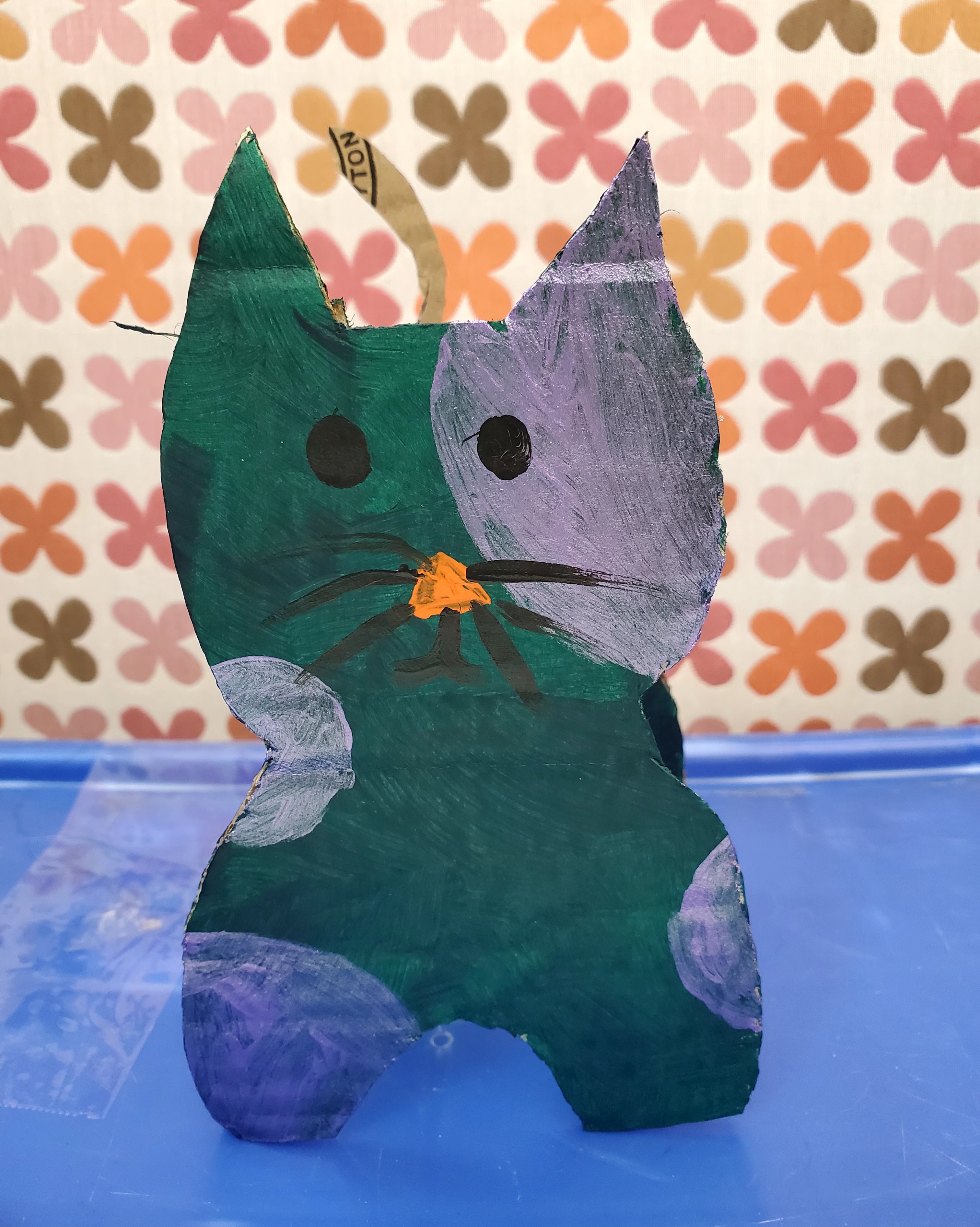 Green cardboard cat with purple splotches, orange nose, black whiskers, black eyes, sitting on top of a blue box with a background of flowers in shades of orange, pink, brown, and yellow