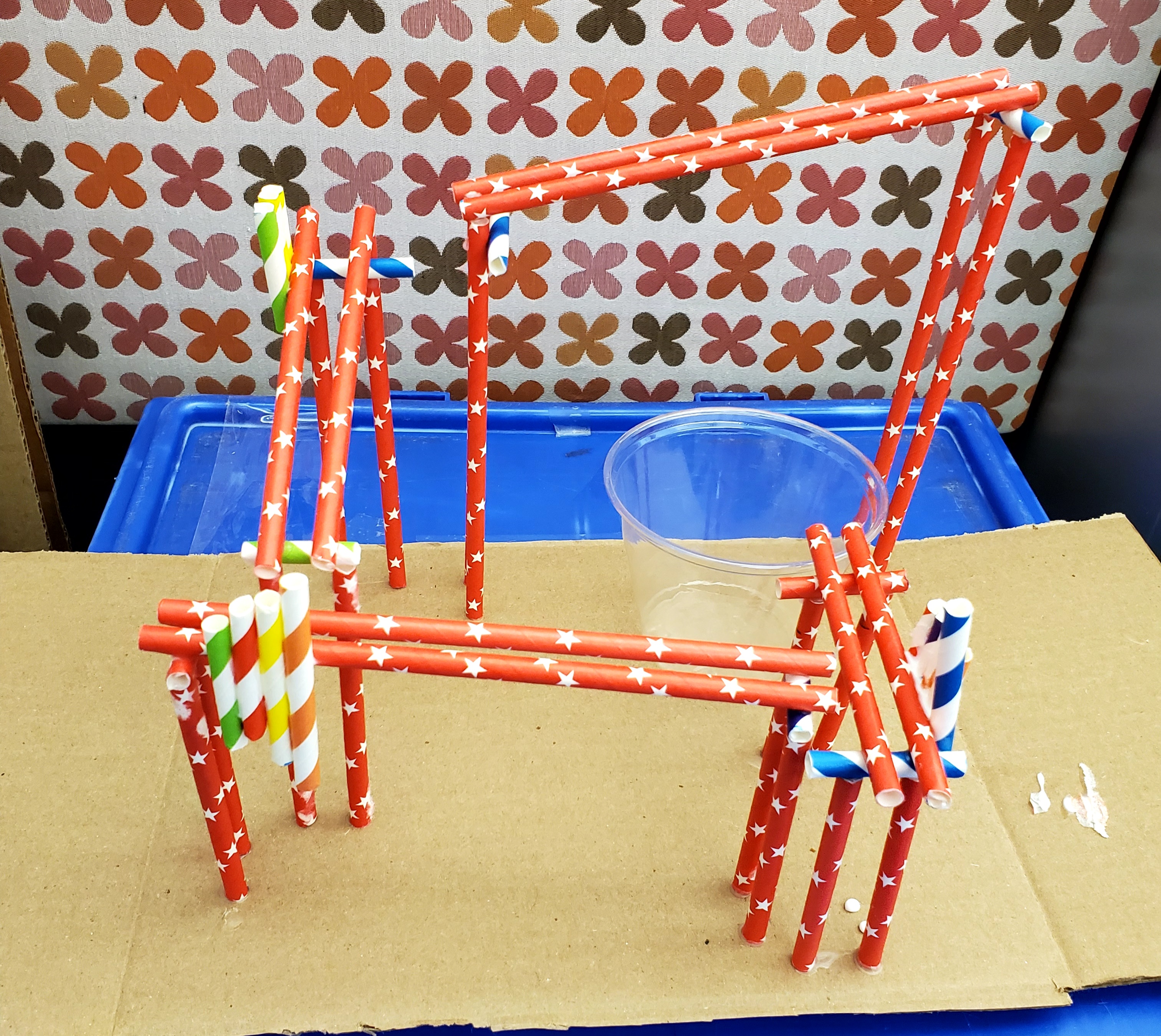 Paper straw rollercoaster made of colorful paper straws and a plastic cup all glued to a piece of cardboard sitting on top of a blue box. Behind the rollercoaster is a panel with a design of flowers in shades of orange, brown, and pink over a white background.