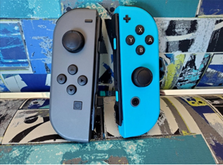Two Nintendo Switch controllers: a grey one to the left, a blue one to the right, on top of a colorful seat background with splotches in shades of blue, tan, and green