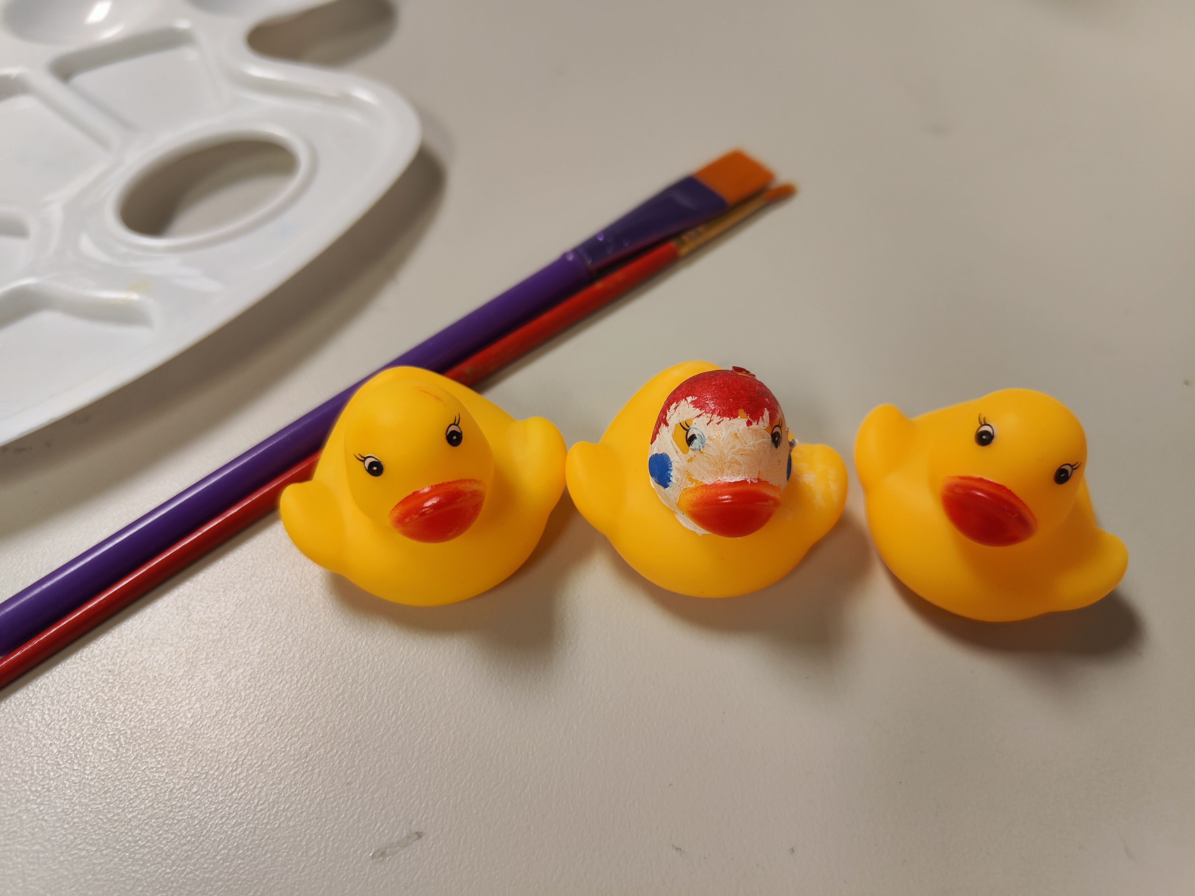 3 yellow rubber ducks with middle duck with clown paint with 2 paintbrushes and paint palette