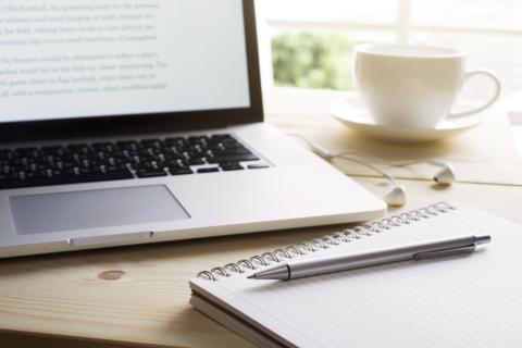 Laptop on desk next to a spiral notebook with a silver pen resting on top