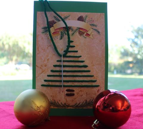 Christmas card with embroidered Christmas tree. Embroidery needle with yarn on it. Two ornaments. Everything laid out on a red cloth.