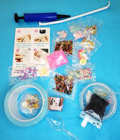 Nano Tape Squishies supplies on a light blue background: Colorful glitter, blue scissors, two Nano Tapes, instruction sheet, straw, colorful slime, and more