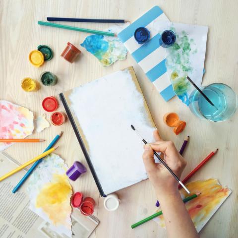 Paints and a notebook. A hand is holding a paintbrush.