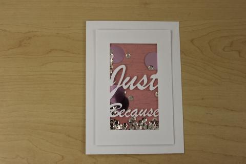 White greeting card with clear window in the middle. The words "just because" are in the window. Sequins and other small pieces are behind the window.