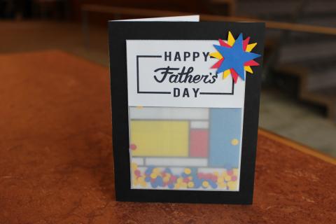 Father's Day shaker greeting card. Red, blue, and yellow confetti inside of a vellum. Father's Day text placed on the outside of the vellum. Black background. Blue, yellow, and red paper star decorations on the front.