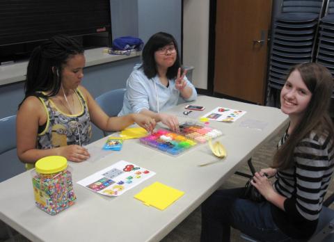 Three teens at a table, two looking at the camera smiling, and making perler bead creations.