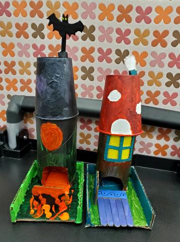 To the left: Scary fairy house with a cauldron bubbling over in the yard. To the right: Cutsey fairy house with a mushroom roof and a purple walkway in the yard