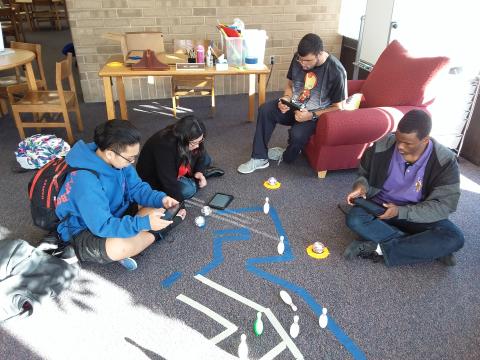 Several teens, some sitting on the floor, use spheros and tablets to figure out a maze puzzle.