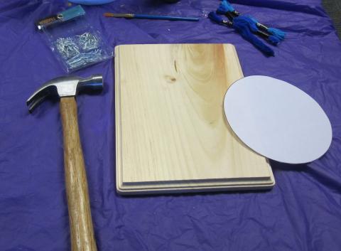 Supplies for String Art on a purple background: hammer, wooden plaque, paper template, nails, and embroidery thread.