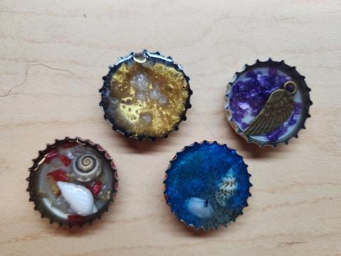 Sample of bottle caps filled with resin and various decorations, such as crushed glass, shells, or metal charm.