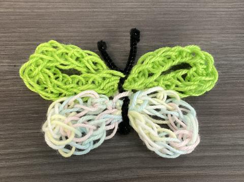 finger knitted butterfly made with green colored yarn, rainbow pastel colored yarn, and black pipe cleaner