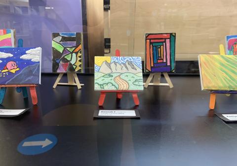 Photo of 5 colorful mini canvases, with easels, decorated with colorful paintings. These canvases are inside of a glass case.