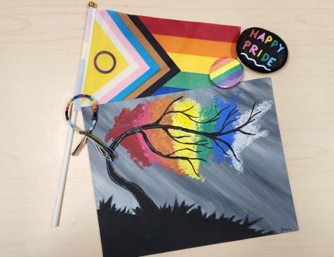 A inclusive pride flag, pride pin, a painted rock saying 'happy pride', and a pride-themed painting