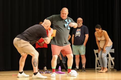 Two men acting out a scene. One is bent over looking through a small cone. The other is holding a toy to his chest. Bystanders observe from the background. One woman in the background is bent over laughing.