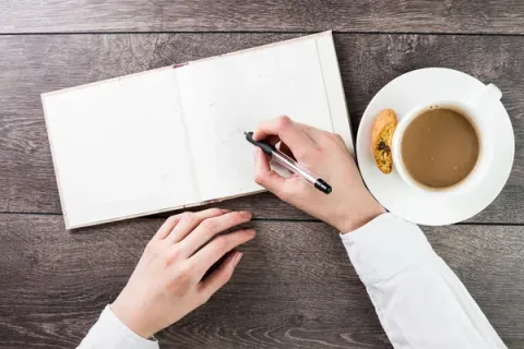 A blank journal sits next to a coffee cup, and a hand holds a pen over the pages.