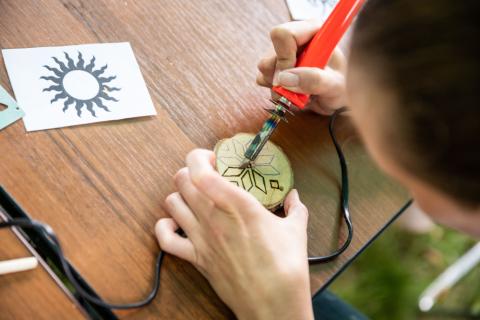 Hands holding a woodburning tool with an orange handle, creating a design in a circular piece of wood. 
