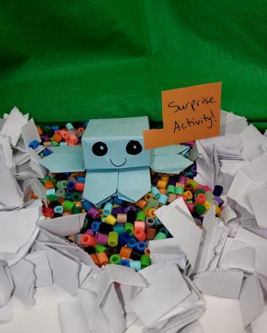 A paper box creature sits surrounded by crafting supplies like perler beads, holding a sign that says Surprise!
