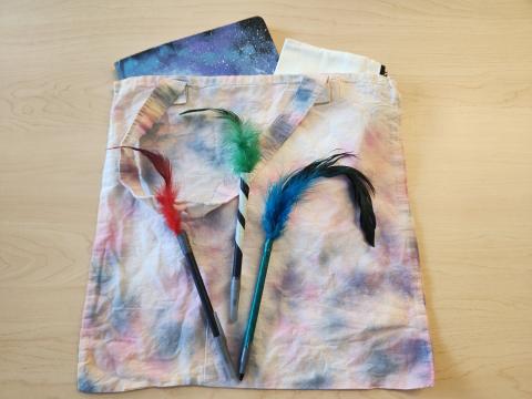 A tie-dyed canvas bag holding a decorated notebook and pencil bag, with a couple pens decorated with feathers and washi tape on top.