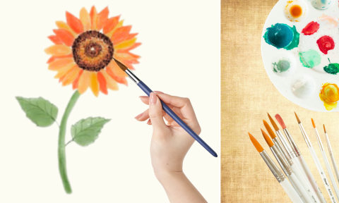 A hand holding a watercolor brush painting a sunflower with a paint palette and more brushes to the side.