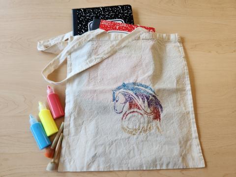 Canvas bag painted with a horse head, holding a black notebook and red notebook, next to three bottles of puffy paint and two paint sponges.