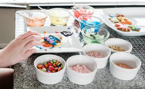 Hand over partially decorated gingerbread cookie on a plate with bowls of colored sprinkles and frosting around