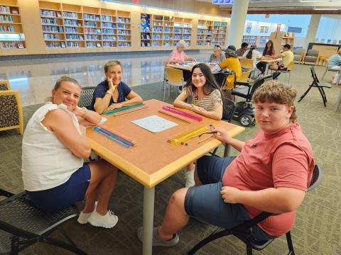 Several tables with adults and teens playing Mahjong. Table in front has a three women and a man looking up from a game