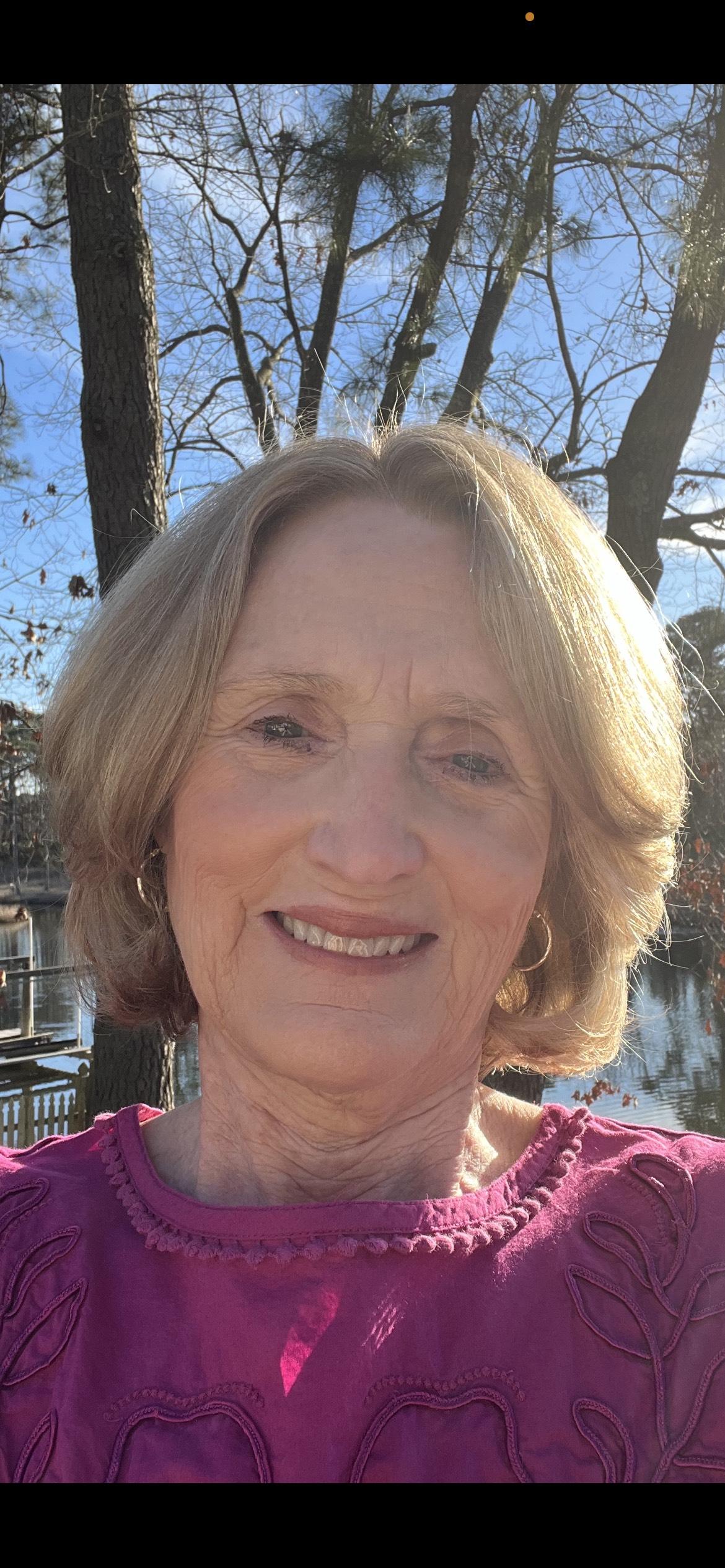 Photo of a smiling woman with blue eyes and short hair wearing a fuschia colored sweater; water and trees are in the background.