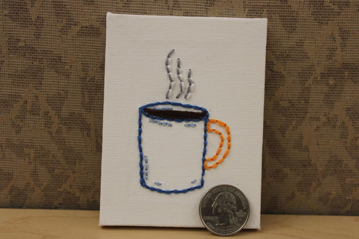 Mug of hot beverage embroidered on a small stretched canvas. Quarter on canvas for size comparison.