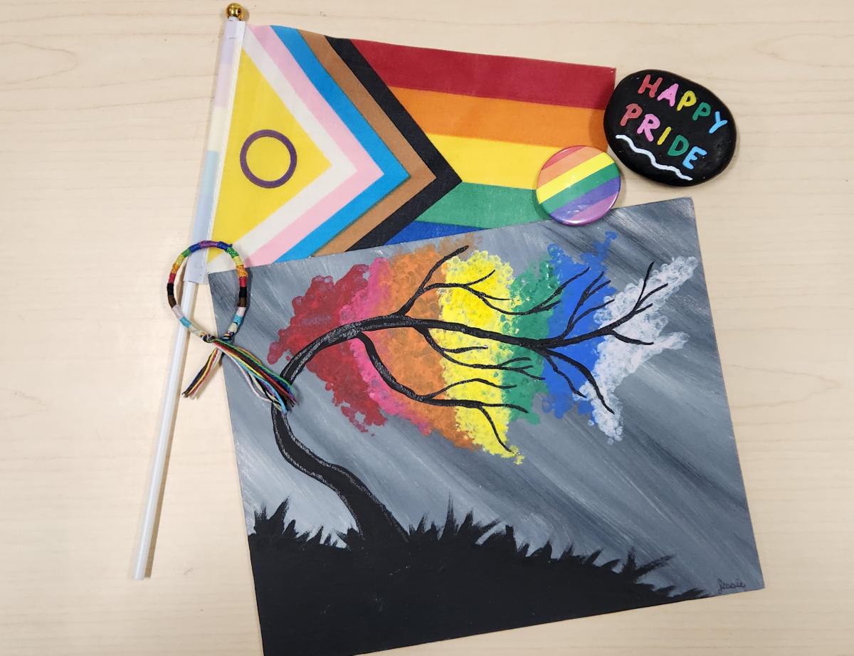 A inclusive pride flag, pride pin, a painted rock saying 'happy pride', and a pride-themed painting