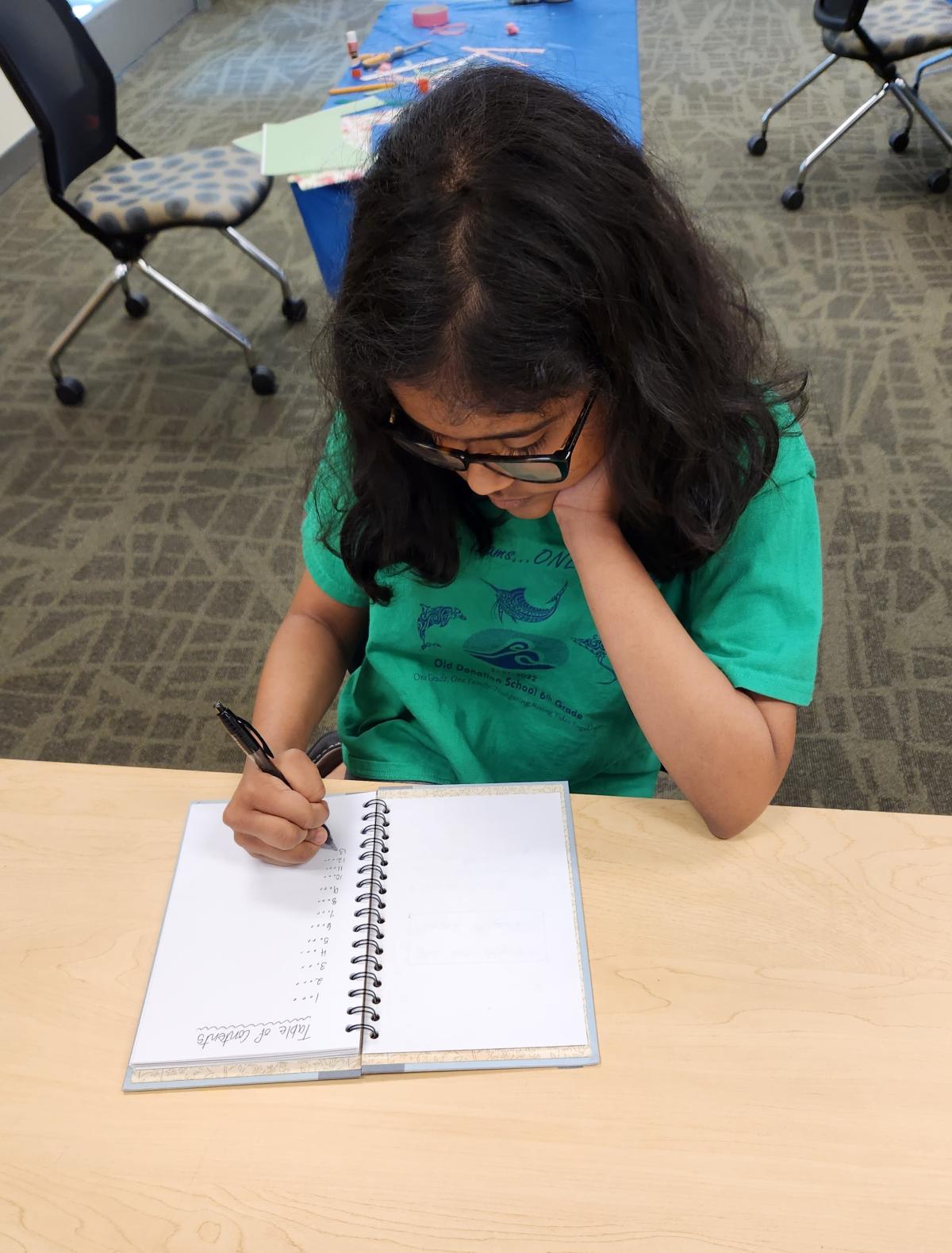 A girl wearing a green shirt is writing in a journal.